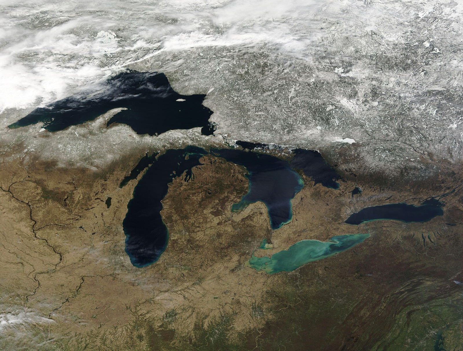 Satellite view of the Great Lakes with the northern land still under snow, the lakes themselves dark open water, and pale green sediment plumes in the western basin of Lake Erie