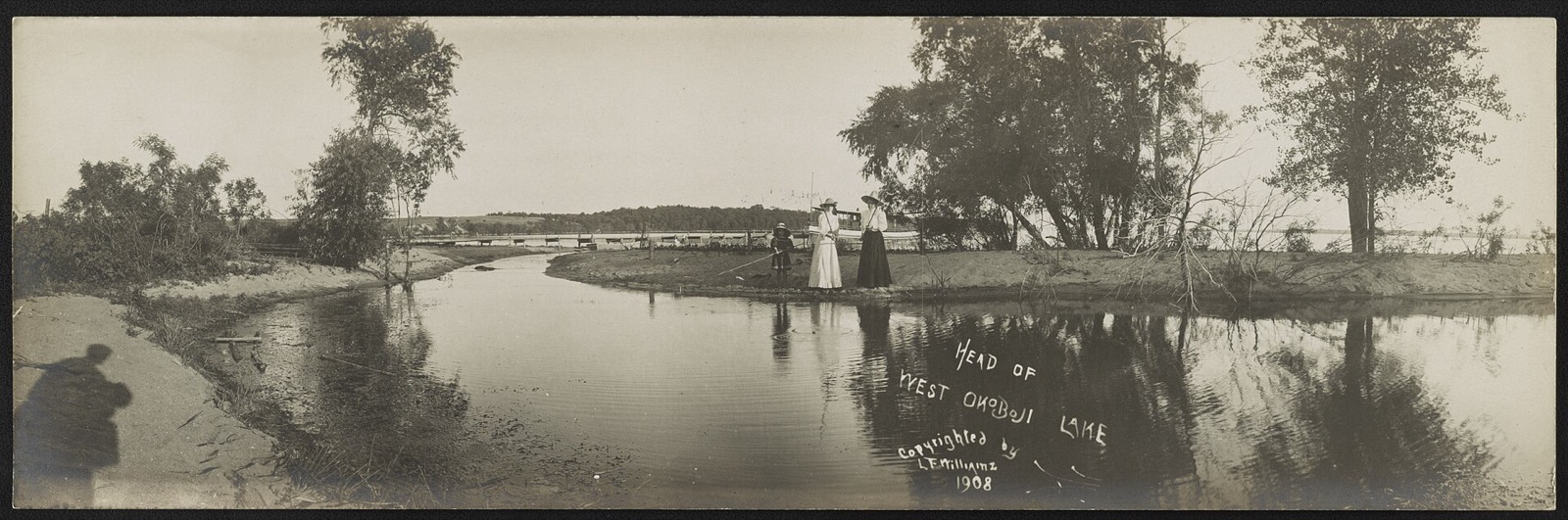 A wide historical panorama of a lake shore with wooded banks, boats and a low pavilion at the water line