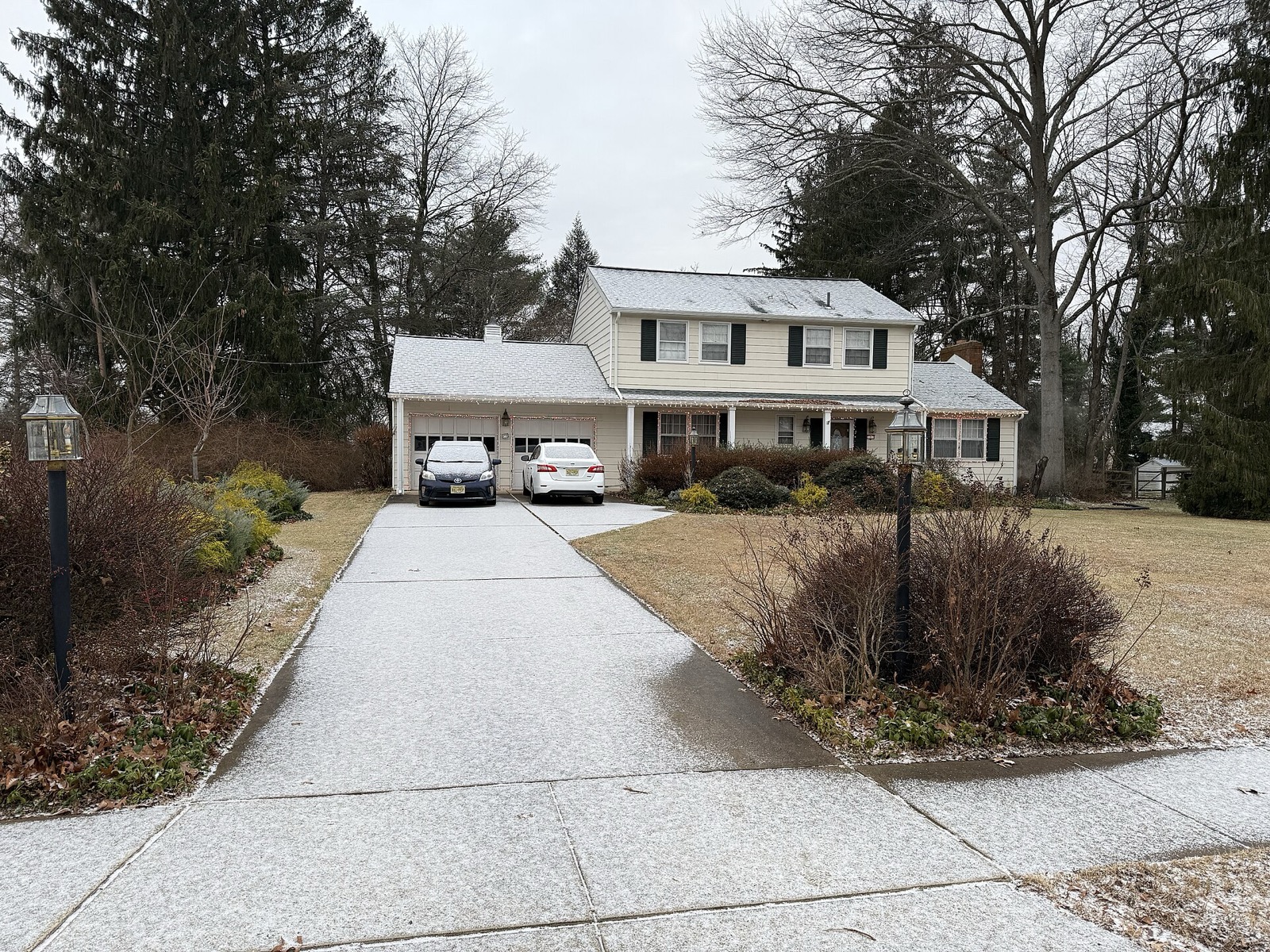 A two storey house behind a long concrete driveway with two cars parked up by the garage doors, a public footway crossing the foreground, and a thin dusting of snow over everything