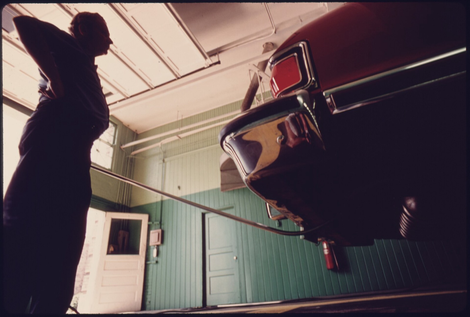 A low angle view inside an inspection garage, a car raised on a lift with a sampling hose run into its tailpipe, and an inspector standing in silhouette against the window watching it