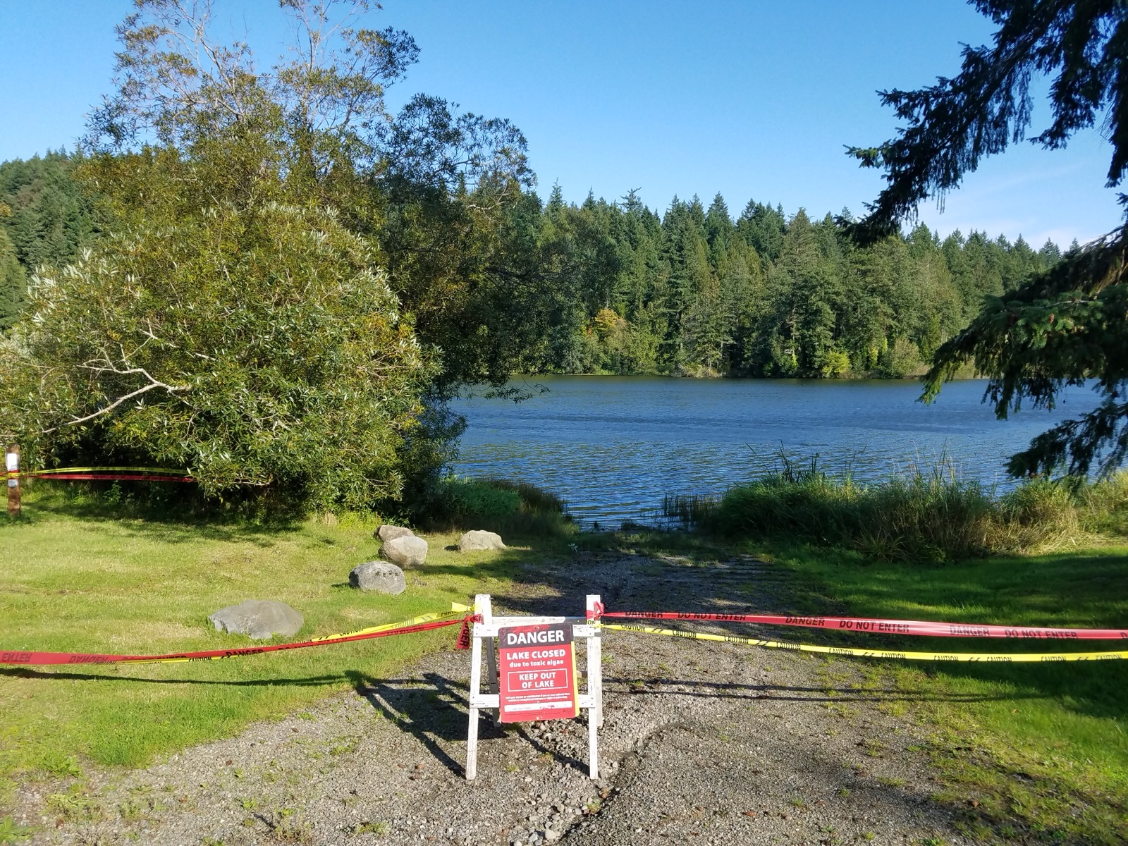 A clear blue forest lake in Washington cordoned off with danger tape and a sign reading Lake Closed due to toxic algae