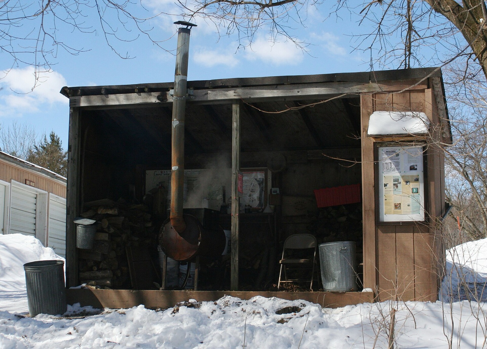 An open-fronted wooden sugar shanty with a stovepipe, stacked firewood and snow on the ground