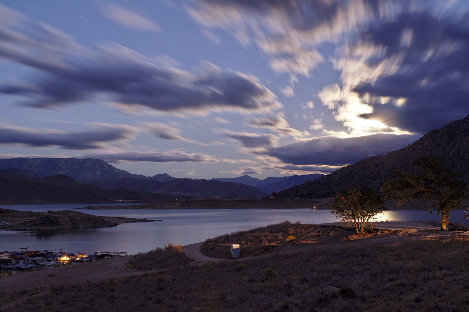 A long exposure of moonlight on streaking clouds over Lake Isabella, with dark mountains and trees along the shore