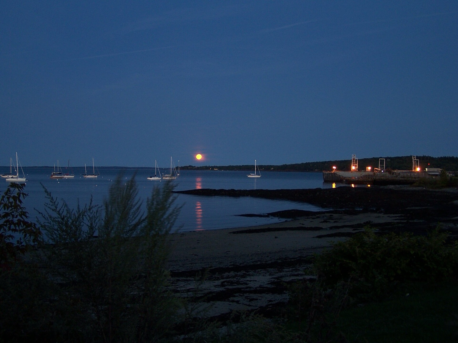 A small orange moon above Rockland Harbor in Maine, with sailboats at their moorings and the moon reflected on the water