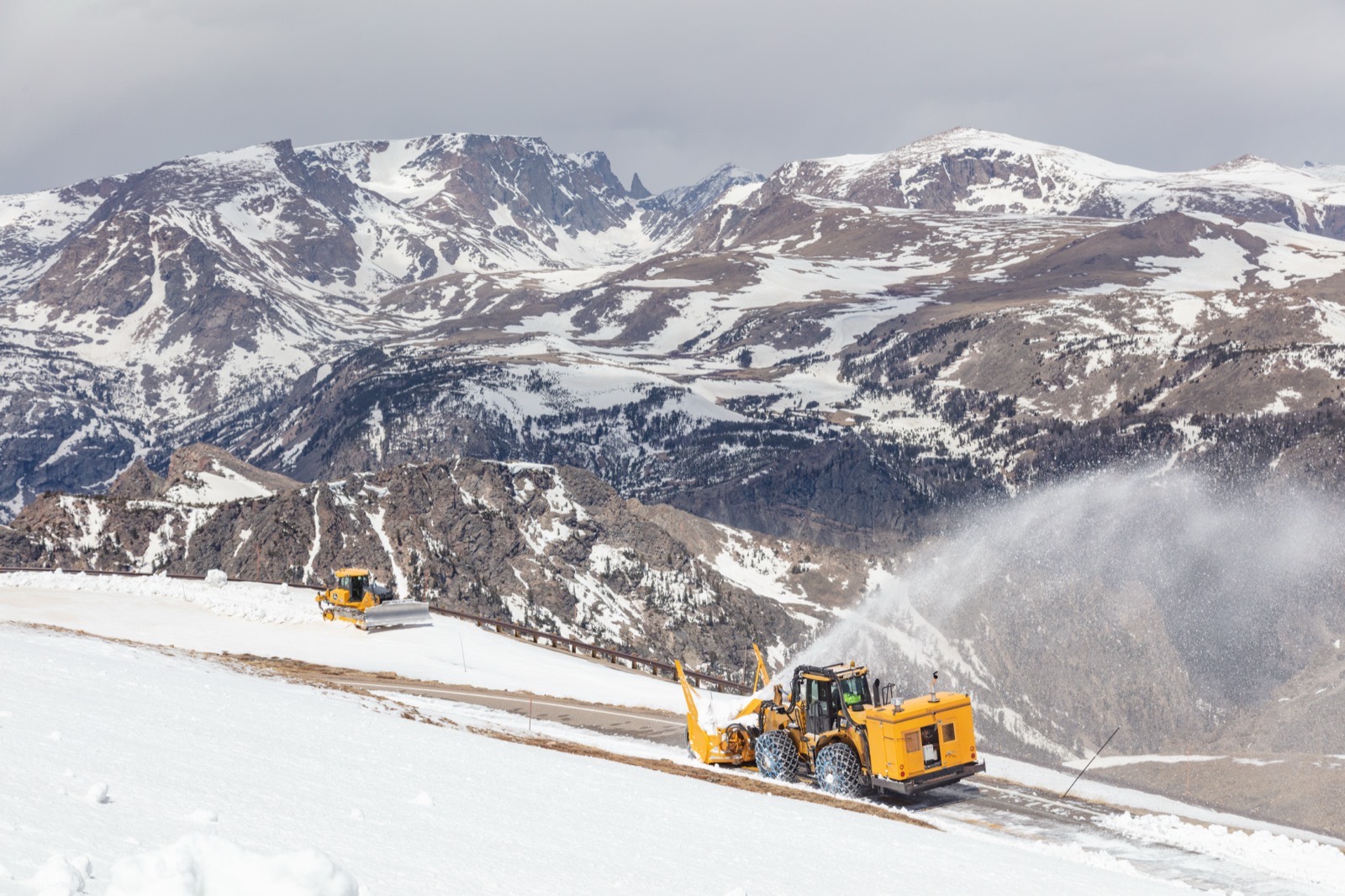 A rotary snowblower throwing a plume of snow off the Beartooth Highway with snow-covered peaks behind, and a second plow working further along the road