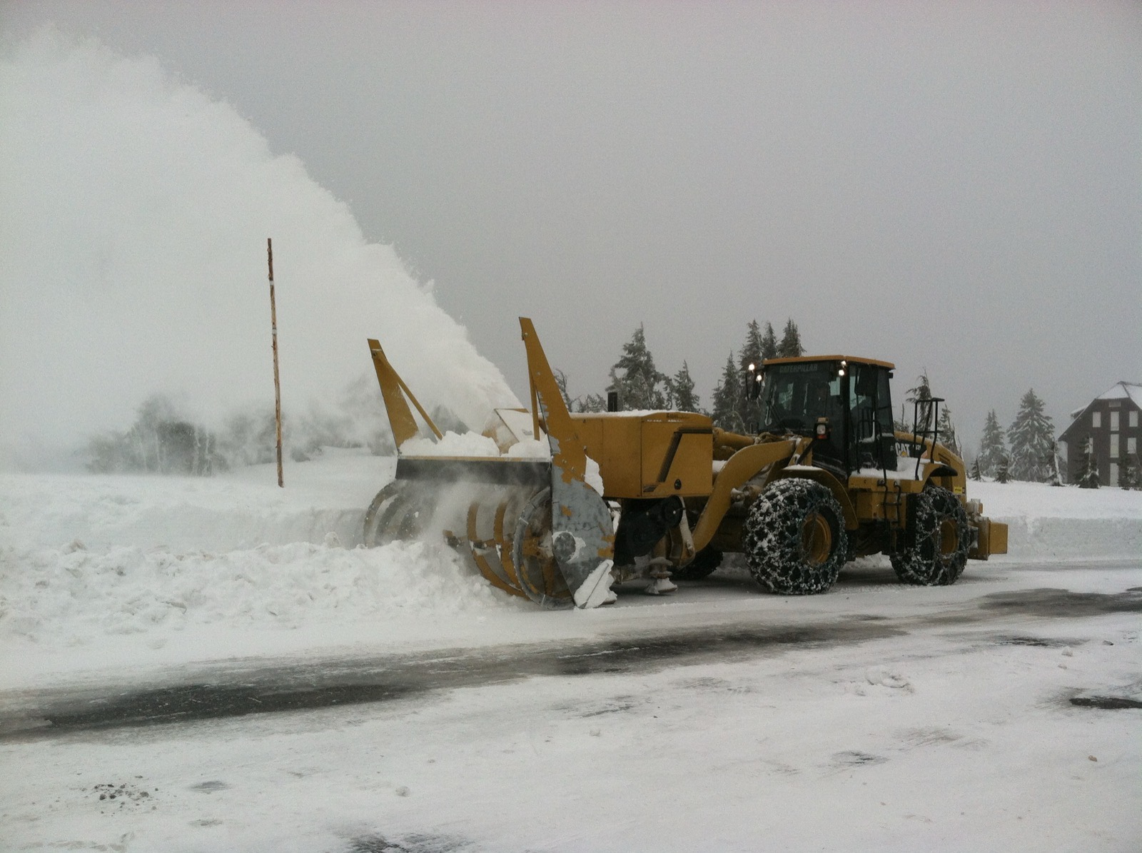 A rotary snowblower throwing a plume of snow beside a plowed road on the rim at Crater Lake, with the lodge behind in falling snow
