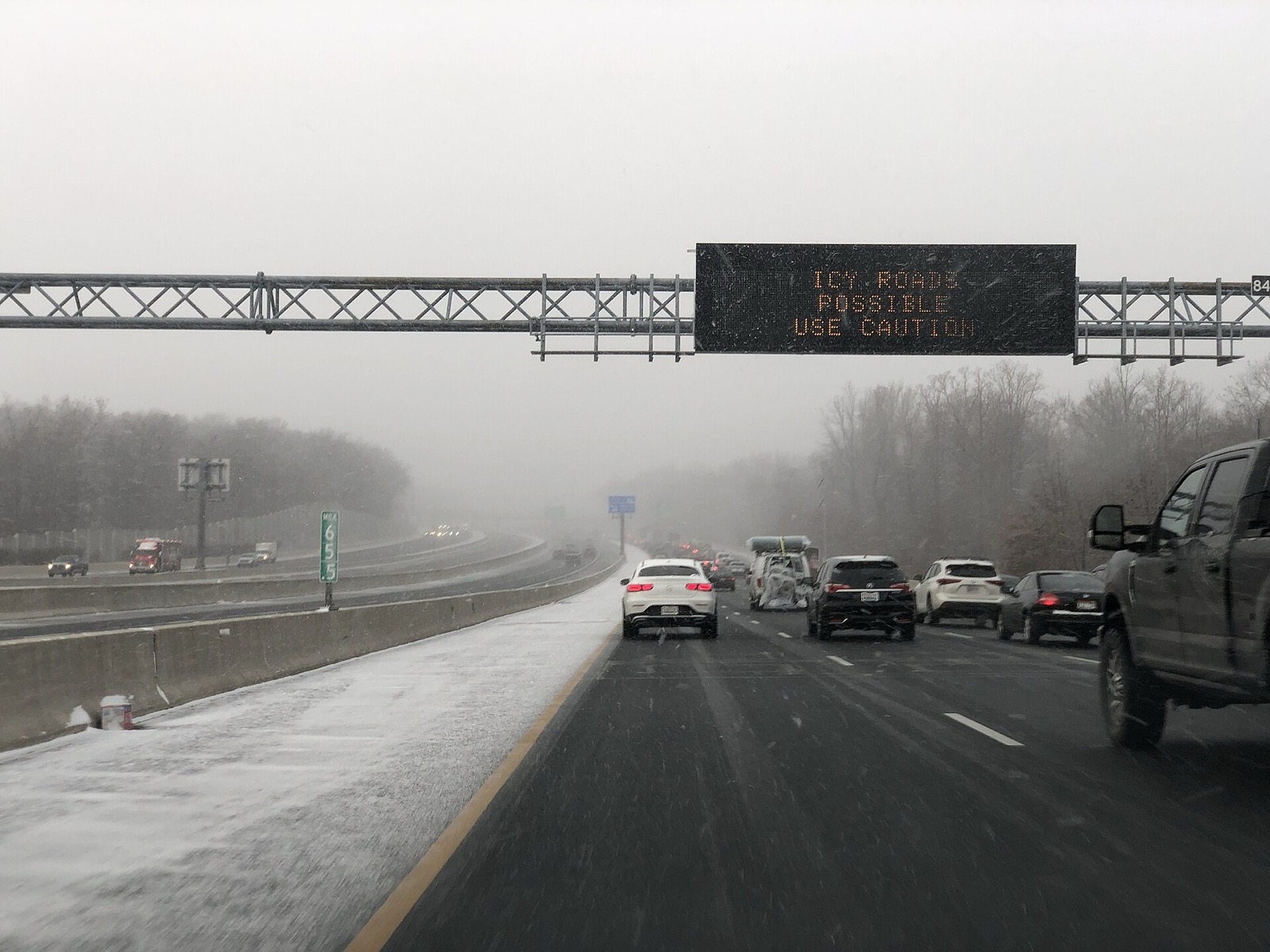 An overhead message sign on a snowy interstate reading ICY ROADS POSSIBLE USE CAUTION, with traffic in the right lanes and a snow covered carriageway beyond the barrier