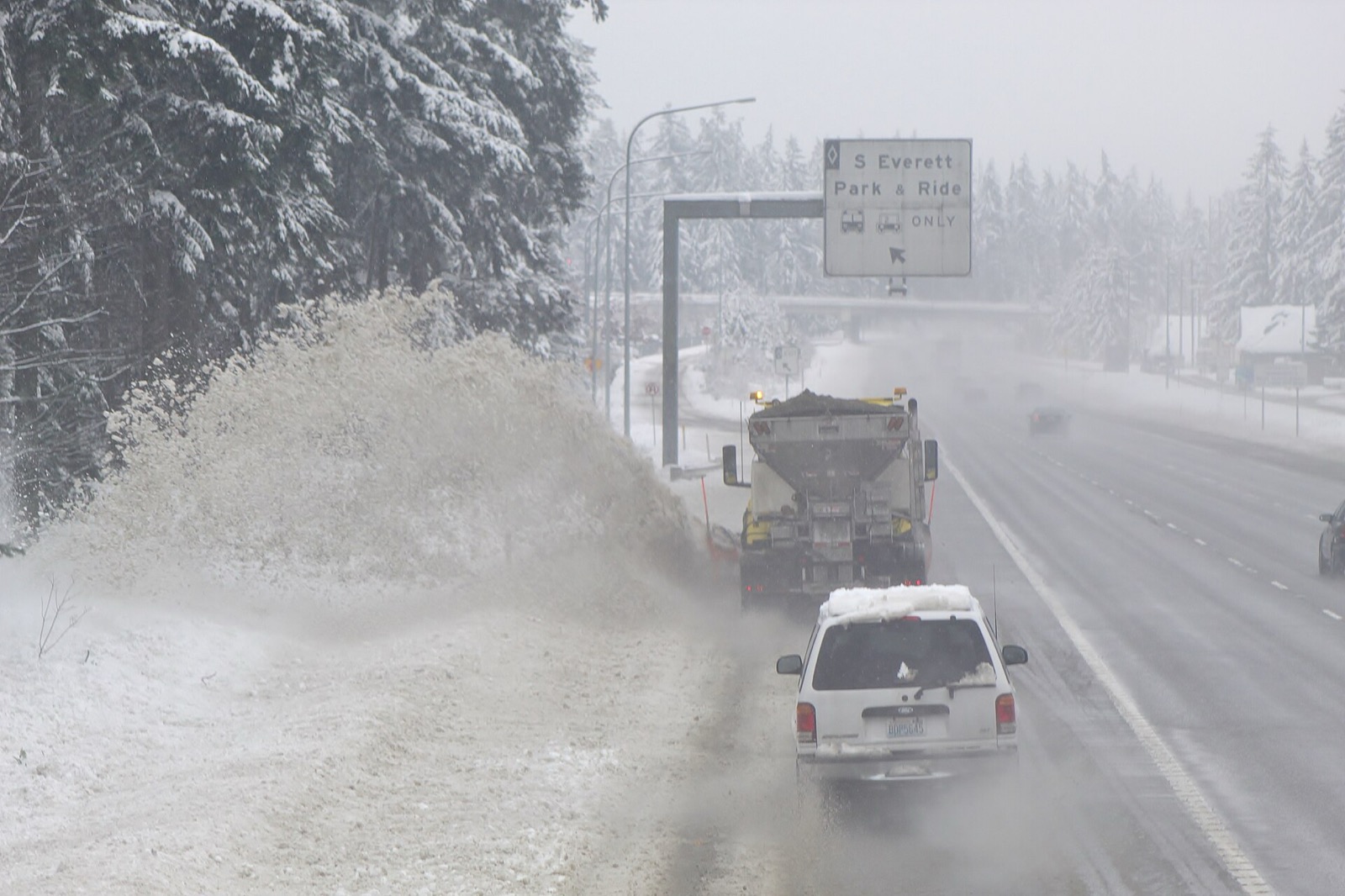 A plow truck seen from behind in heavy snowfall casting a wide plume of snow off the left shoulder of an interstate, with one dark treated lane running away to the right