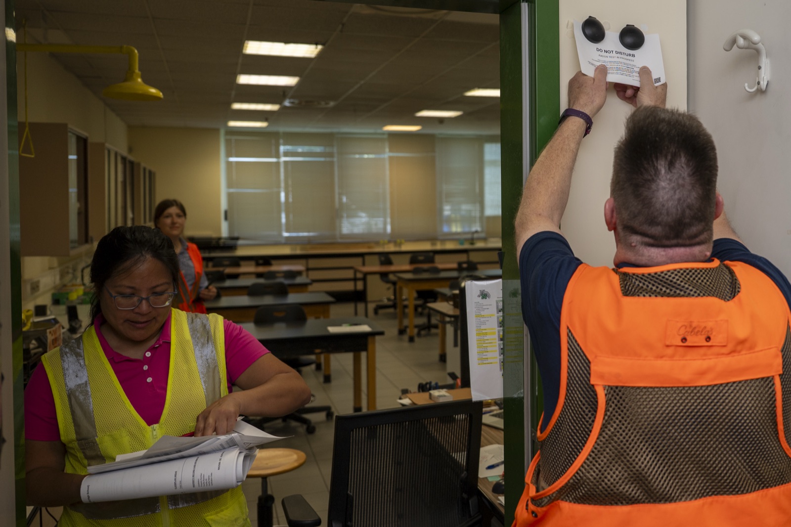 A technician in a safety vest retrieving two dome shaped radon detectors mounted on a do not disturb card on a wall