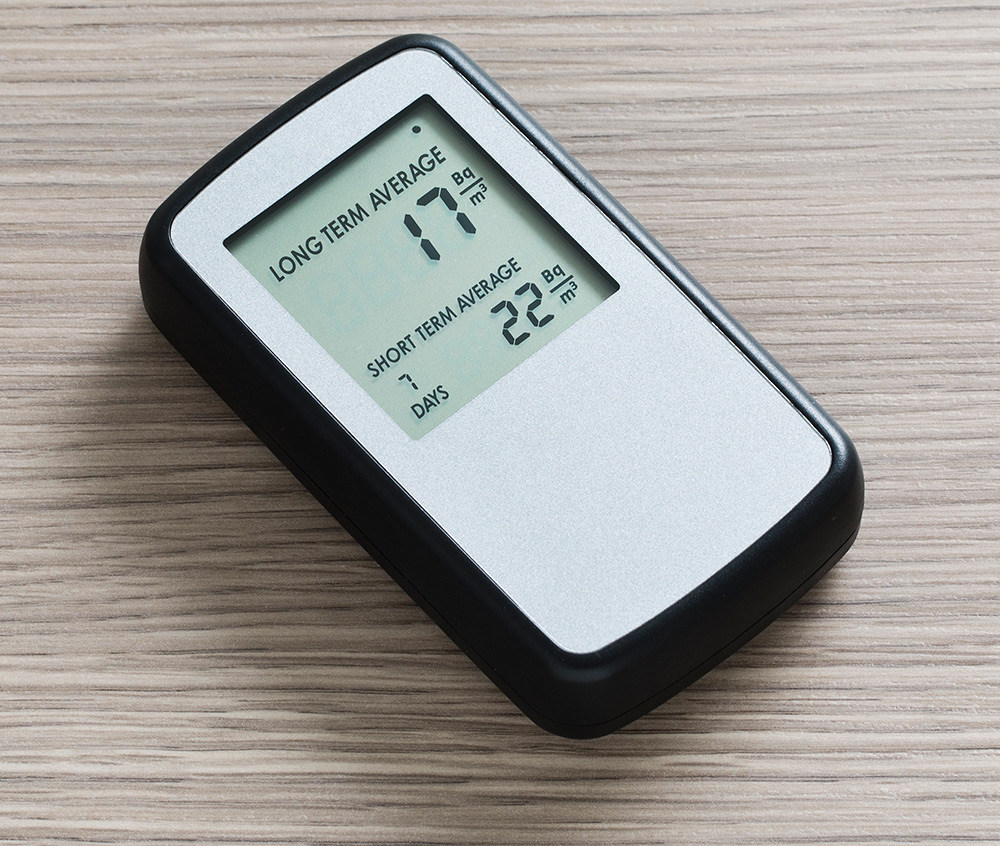A consumer digital radon monitor on a wooden table showing a long term average of 17 and a short term average of 22 becquerels per cubic meter