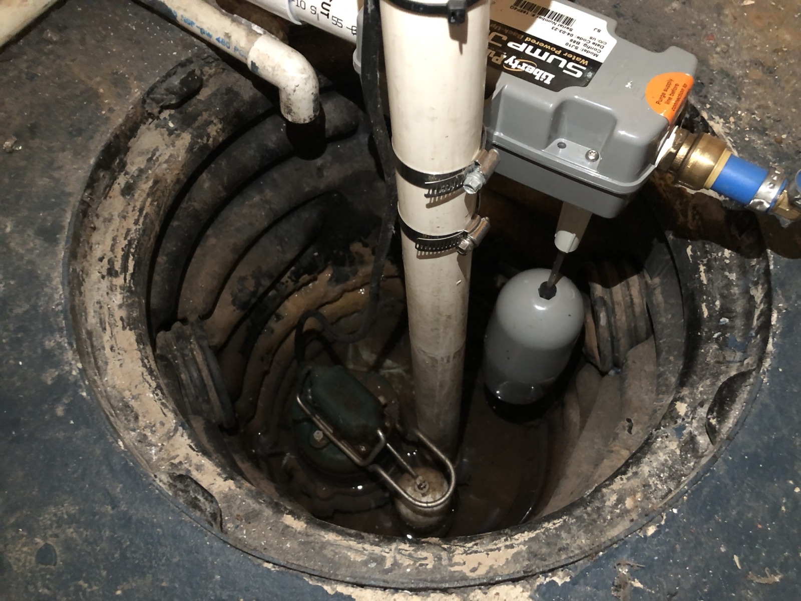 An open sump pit in a basement floor with a sump pump, float switch and discharge pipes visible