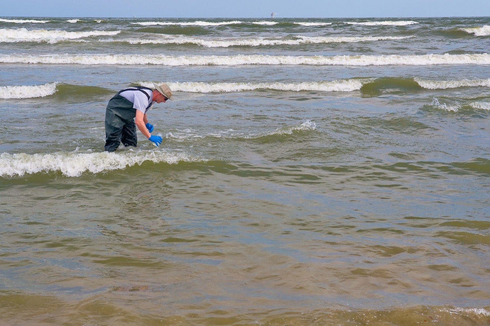 A man in chest waders standing knee deep in the surf, filling a sample bottle with seawater
