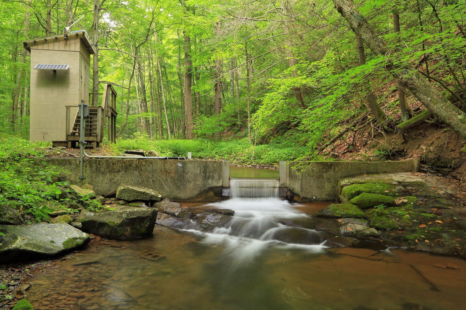 A small concrete streamgage station with a solar panel beside a forest creek and weir