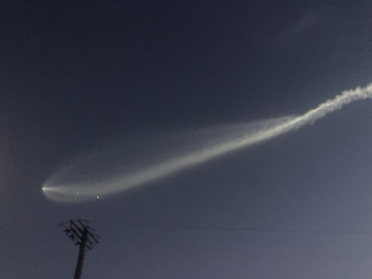 A thin pale trail of rocket exhaust curving across a dark blue evening sky above a power pole, with a small bright point of light at its left end