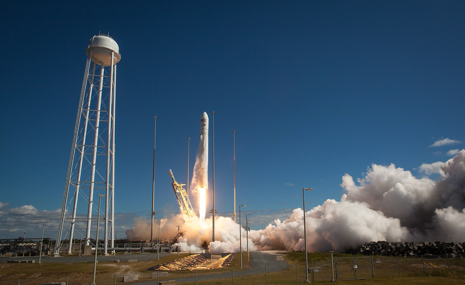 A white Antares rocket lifting off from a launch pad in bright daylight under a deep blue sky, with a tall white water tower at left and a billowing cloud of white exhaust at right