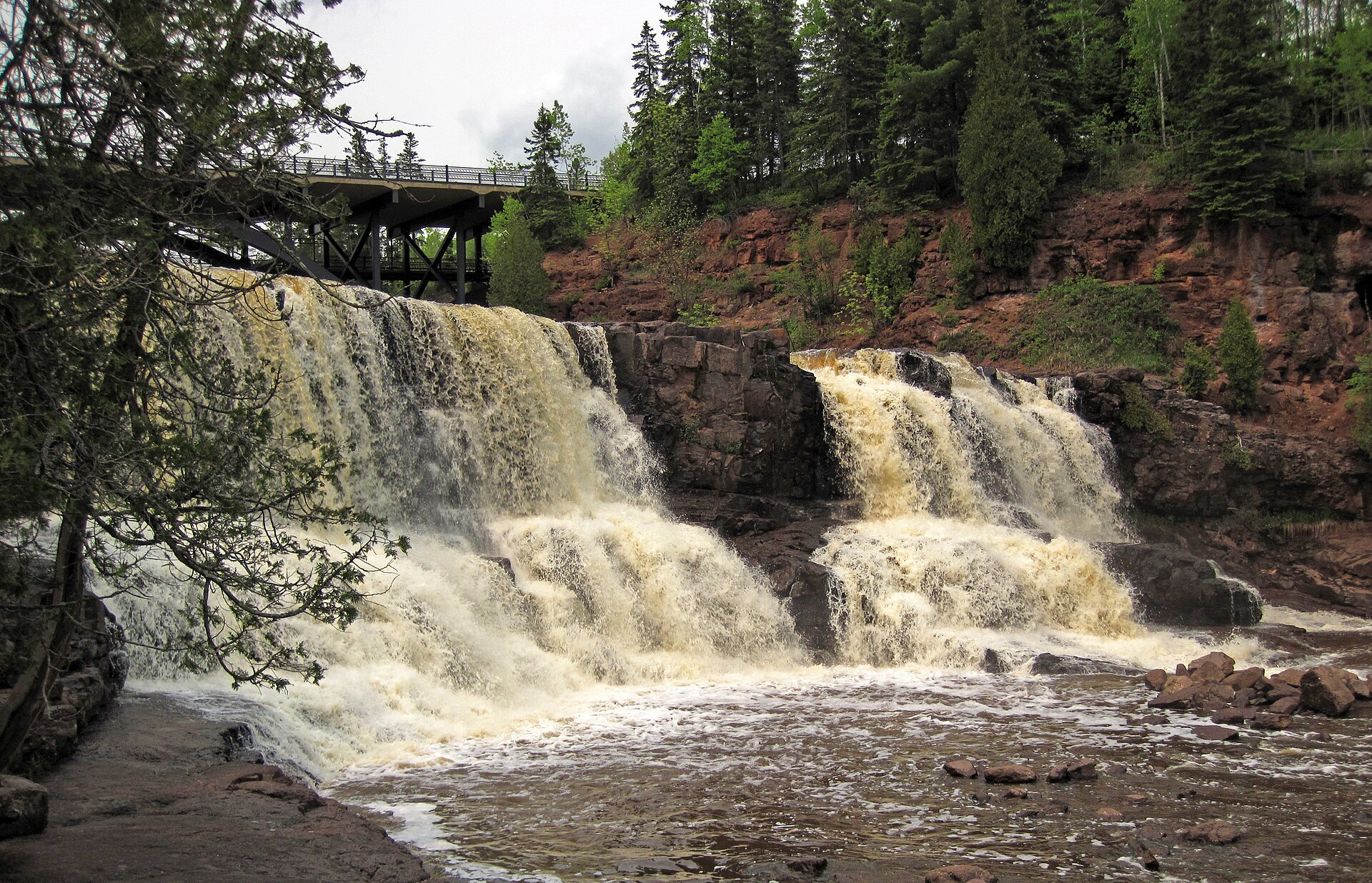 A tea coloured river in high water pouring over two wide rock ledges below a road bridge, in a conifer and birch forest