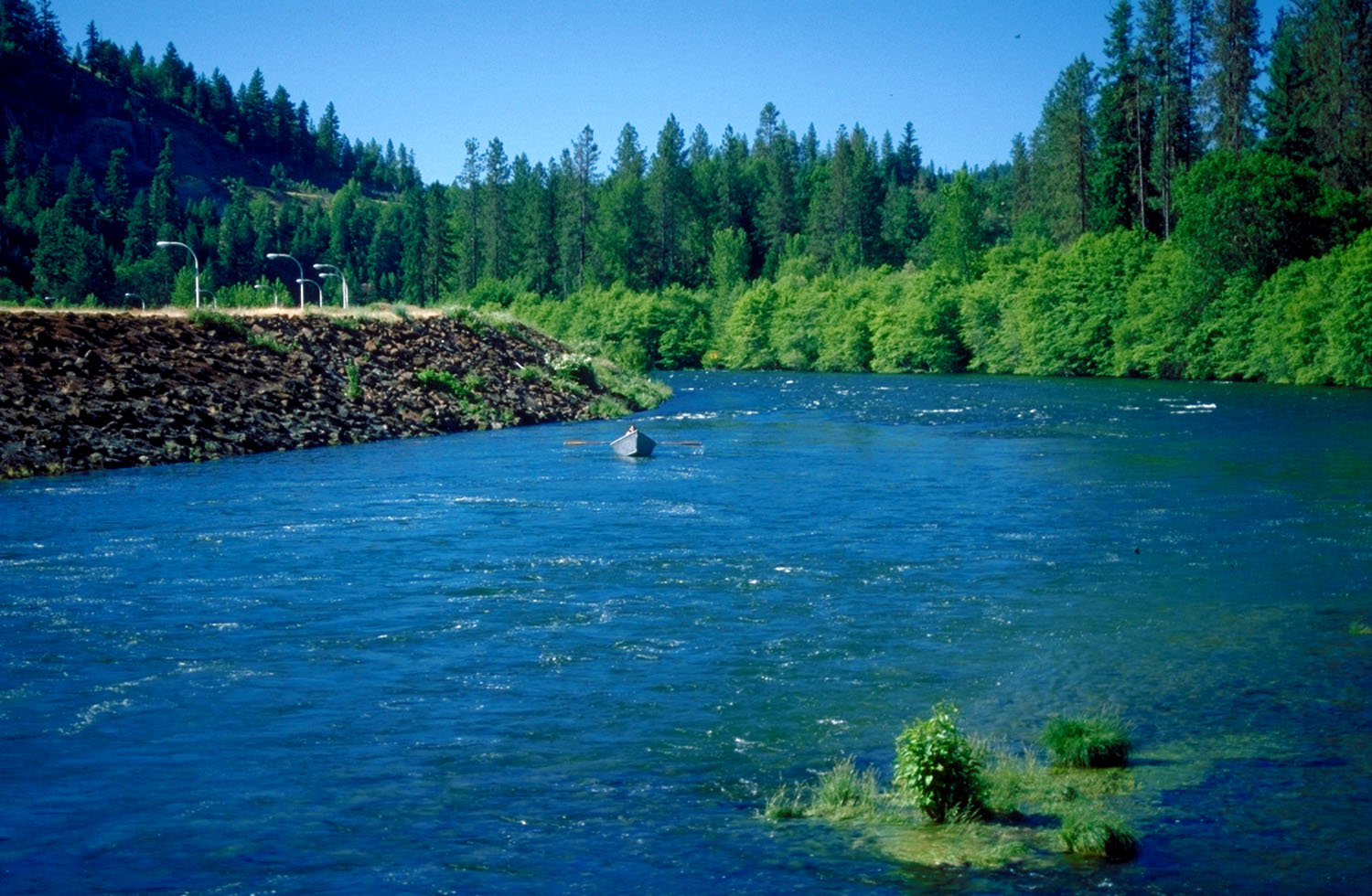 A broad blue green river running fast between conifer forested banks, with a single small drift boat mid channel