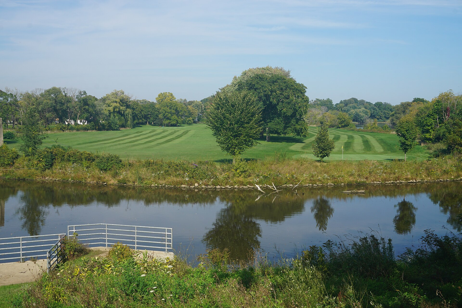A slow river running past a mown riverside park under a clear blue sky