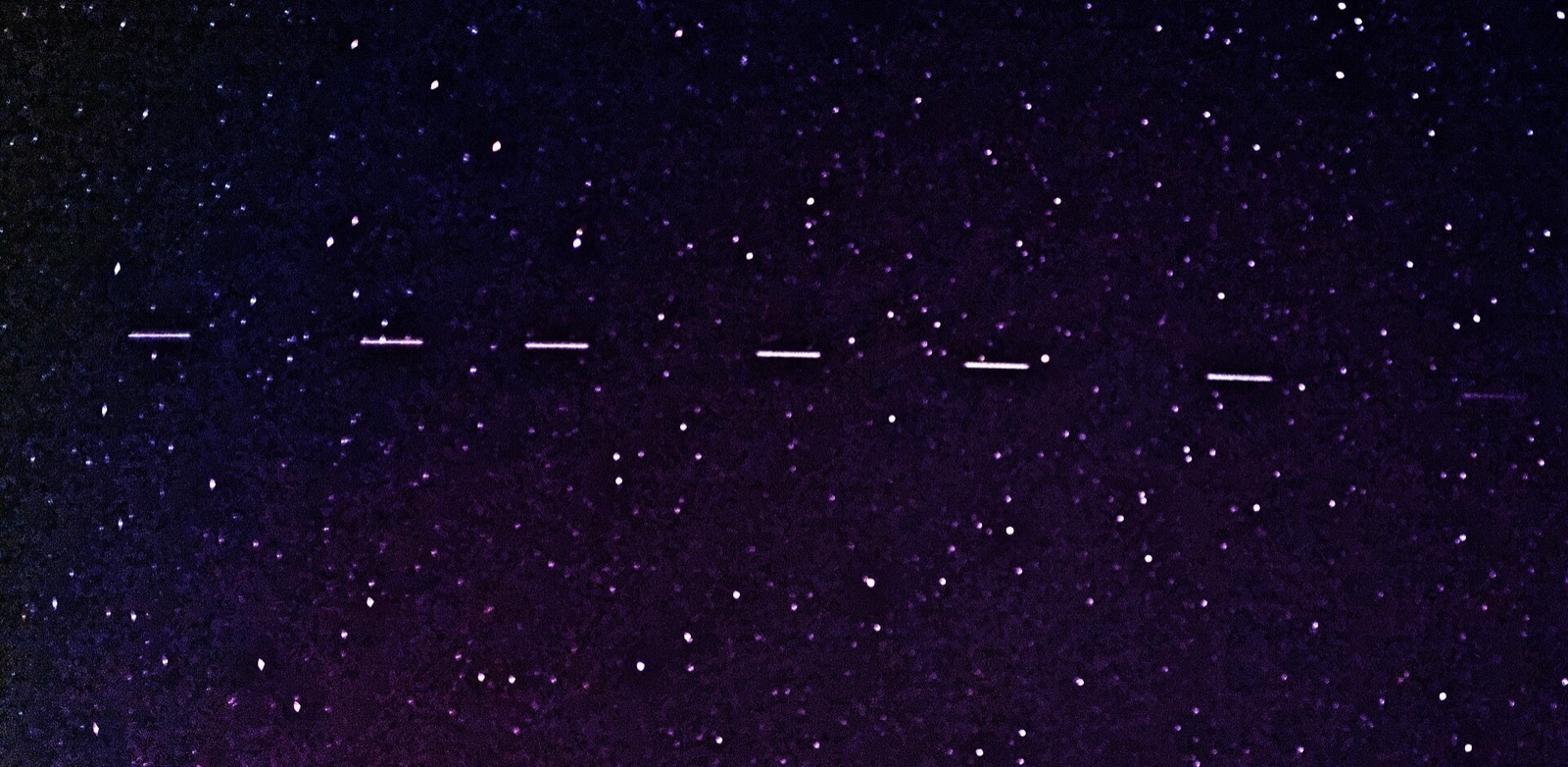 Six short parallel dashes of light in a row across a purple, star filled night sky, each one a satellite moving during a two second exposure
