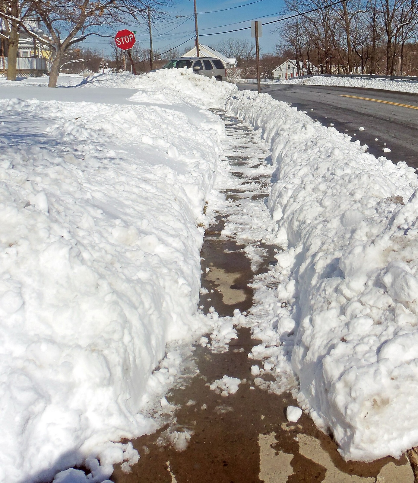 A narrow trench, hardly wider than a boot, cut the length of a sidewalk through snow banked higher than the walk on both sides, a plowed road on one side and a stop sign at the corner beyond