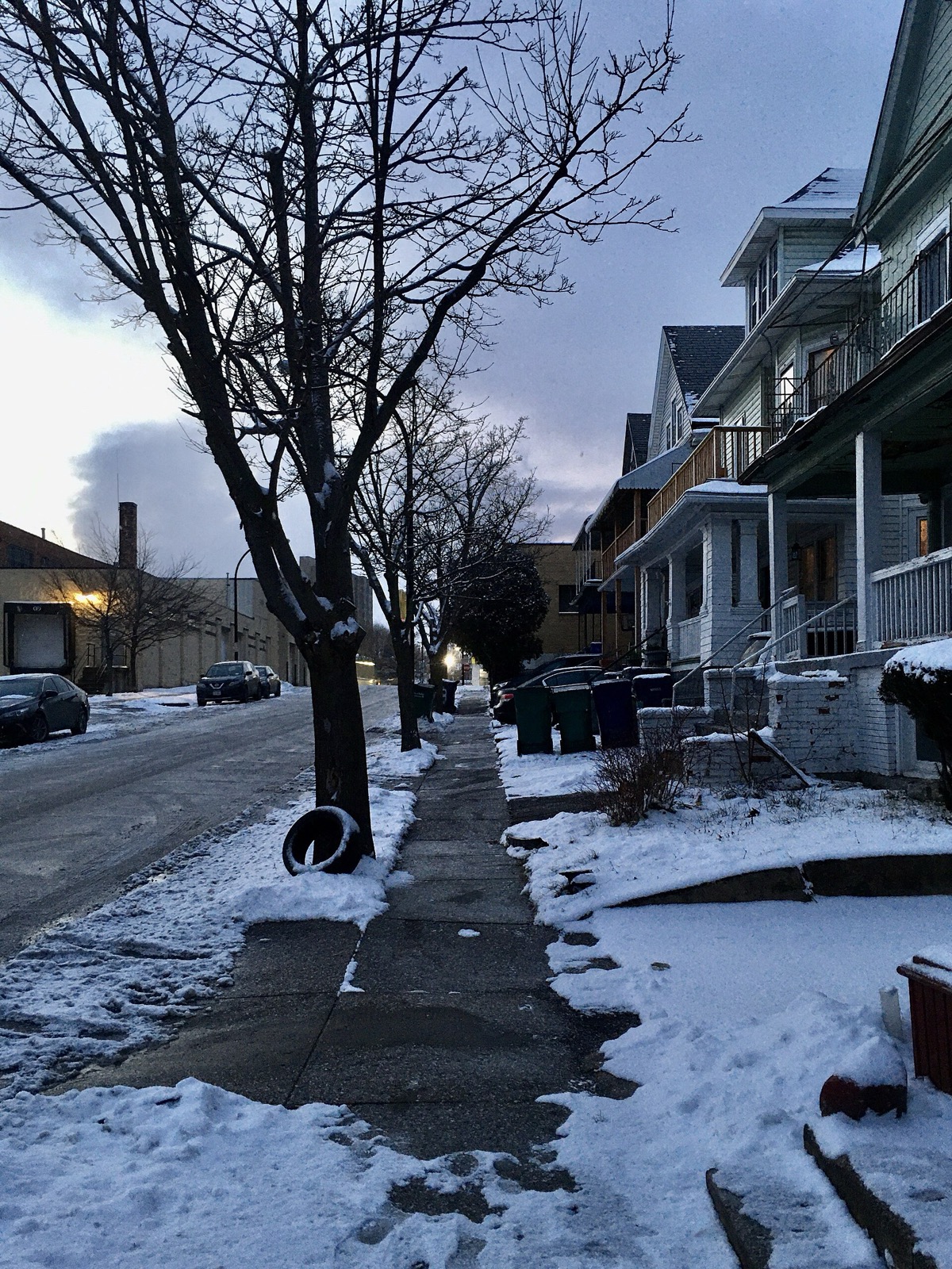 A residential street at dusk in winter, the sidewalk running clear of snow in front of a row of porched wooden houses, wheeled bins out at the kerb and a light snow cover on the lawns either side