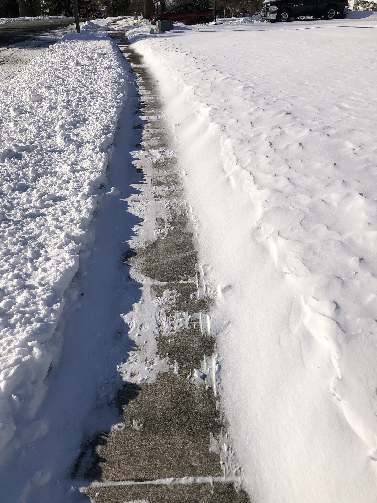 A sidewalk cleared earlier in the day with fresh wind blown snow already lying back across the outer half of it, a plowed road to one side and an unbroken snow field to the other