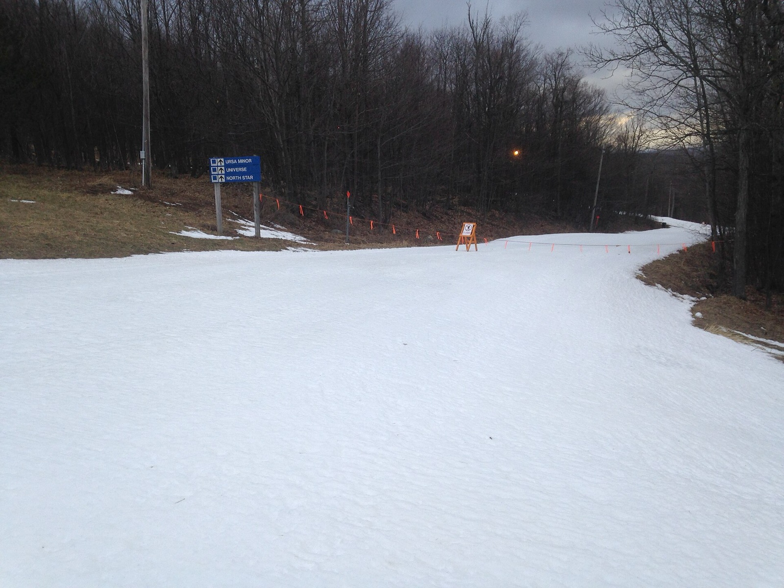 A single narrow strip of manmade snow on an otherwise bare ski trail, with closed-trail rope and signs