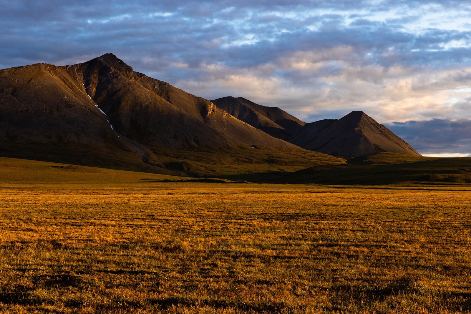 Low golden light across arctic tundra and a bare mountain ridge in the middle of the night, under a broken cloud deck