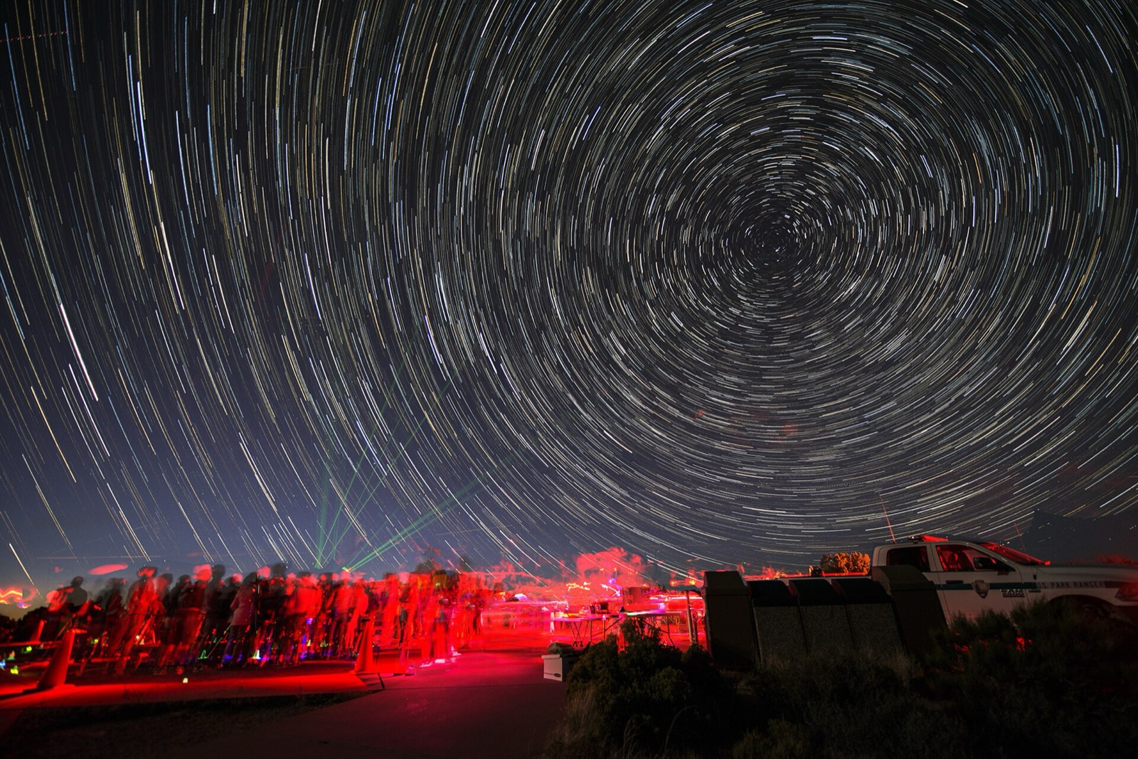 A crowd of people with telescopes under red headlamp light, beneath a long exposure of star trails circling the north celestial pole