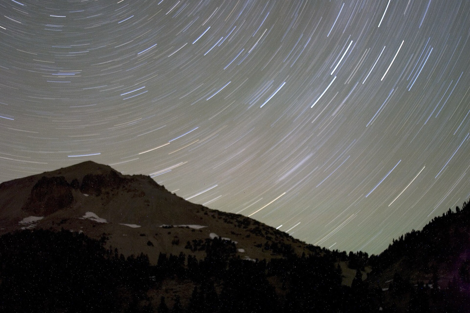 A long exposure of star trails wheeling around the north celestial pole above a snow patched volcanic peak and a dark treeline
