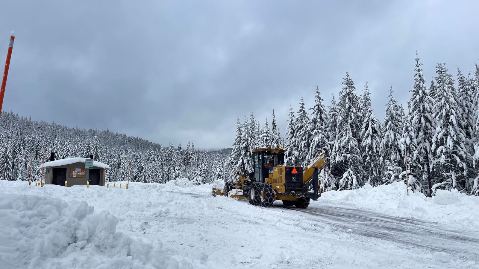 A yellow road grader with tyre chains clearing deep snow from a forest car park beside a small concrete restroom building carrying a green permit required sign, under a low grey sky with heavily laden conifers behind