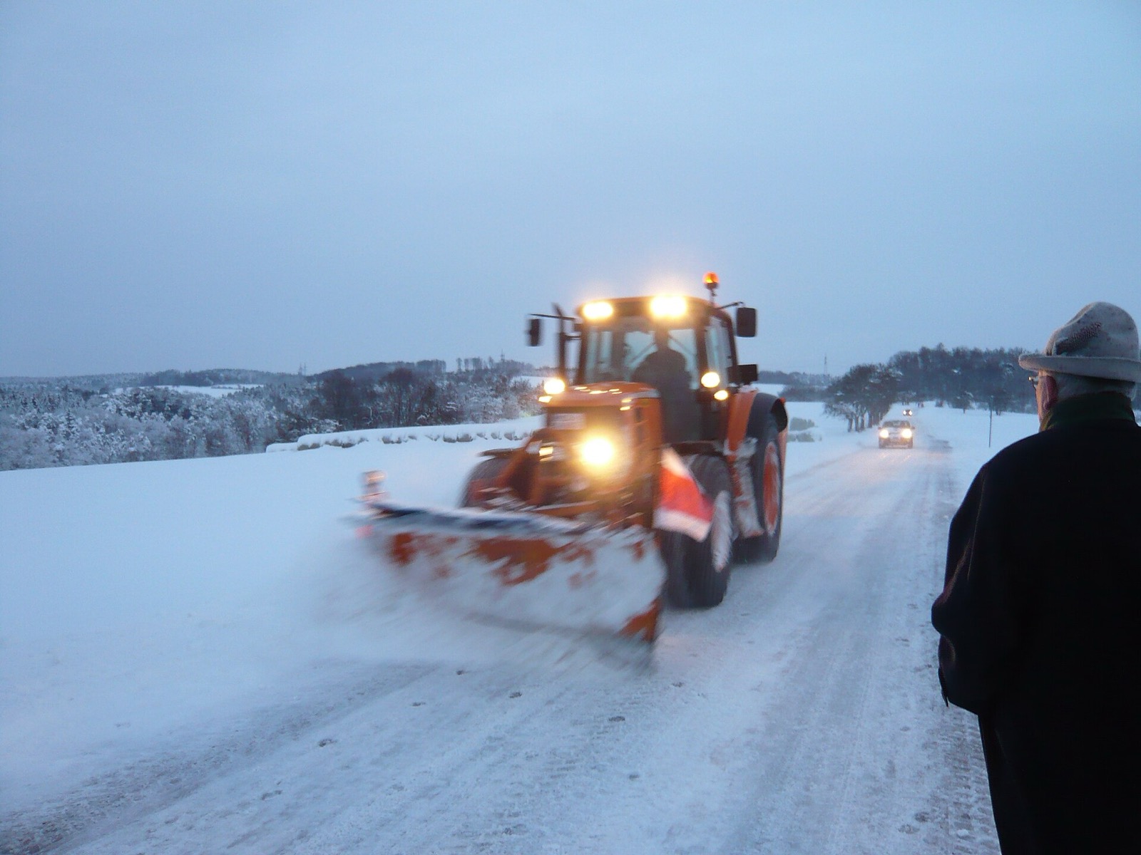 An orange tractor with a front plow blade and its lights on clears a snow covered country road at dawn, a man in a hat watching from the roadside