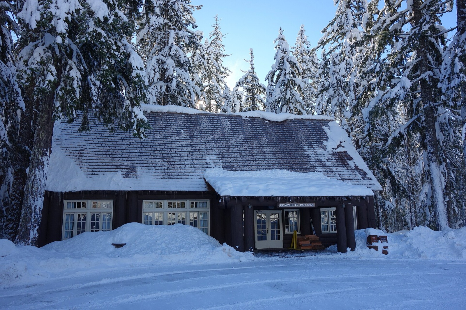 A long single storey log building with a steep shingle roof standing among tall snow covered firs, deep snow lying along the roof and curling over the porch edge in a thick lip hung with icicles, drifts banked to the window sills, and a small carved sign reading COMMUNITY BUILDING above the doors