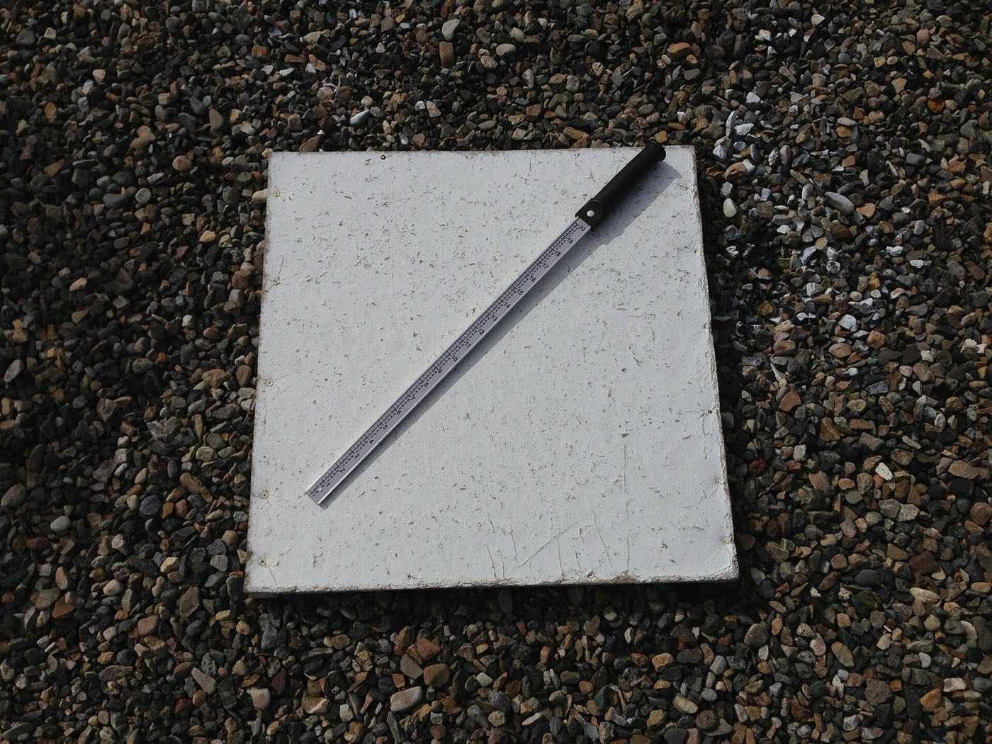 A white square snow board lying on gravel with a metal snow measuring stick laid across it diagonally