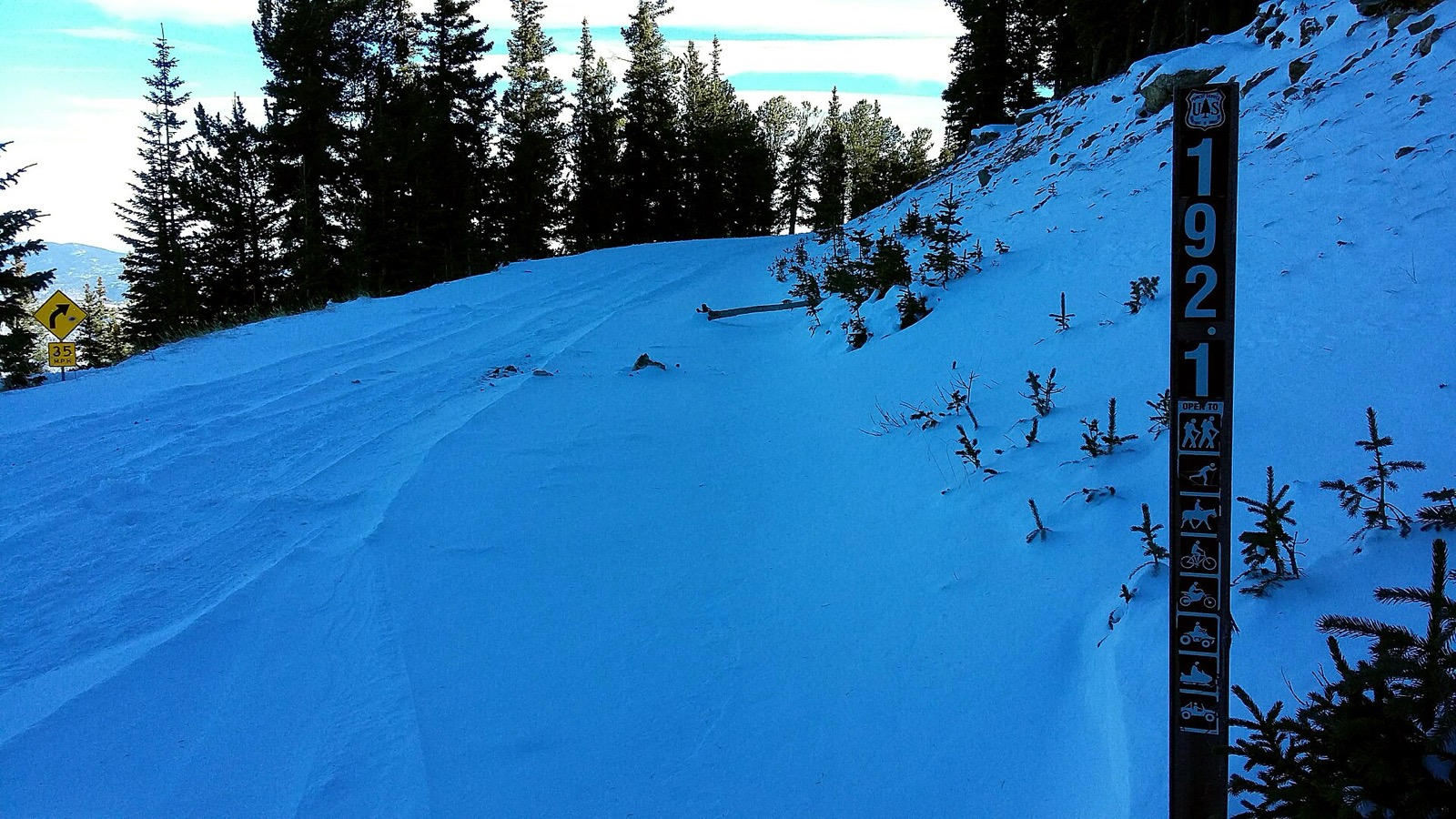 A U.S. Forest Service route marker post numbered 192.1 beside a snow covered forest road, carrying a vertical strip of pictograms showing which uses the route is open to, including a snowmobile