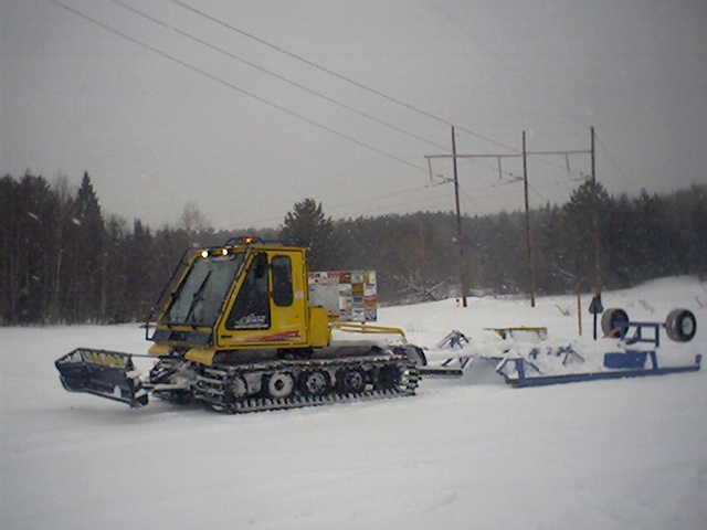 A yellow tracked grooming tractor parked in falling snow with a wide steel drag hitched behind it and a second drag stacked at the edge of the clearing