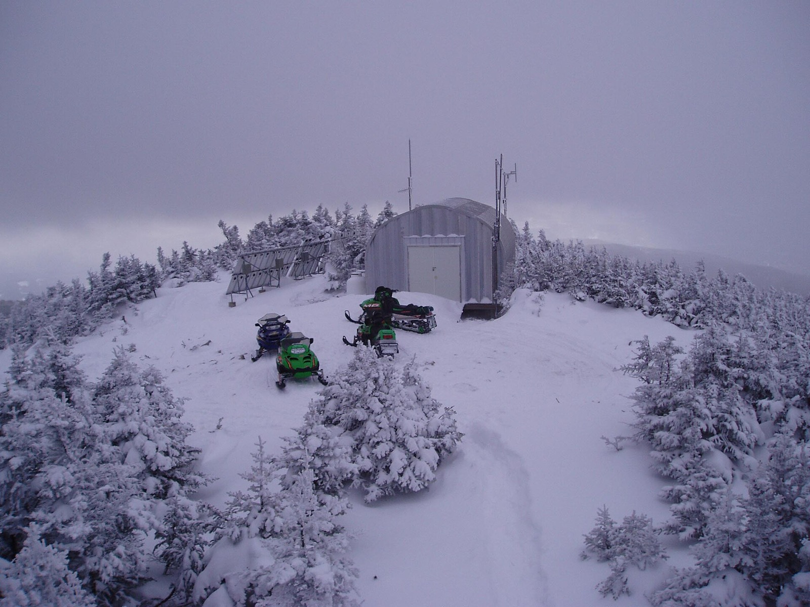 Three snowmobiles parked in fog beside a metal hut and a solar array on a rime covered summit, surrounded by stunted spruce buried in snow