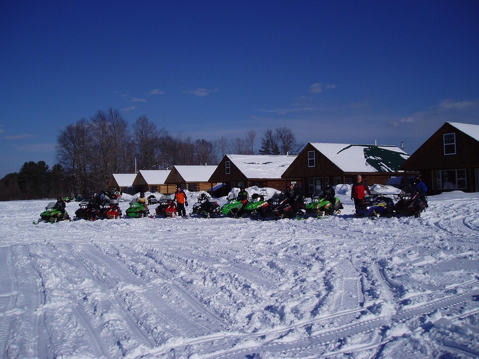 A long row of snowmobiles parked in front of a line of log cabins under a deep blue sky, with heavily tracked snow filling the foreground