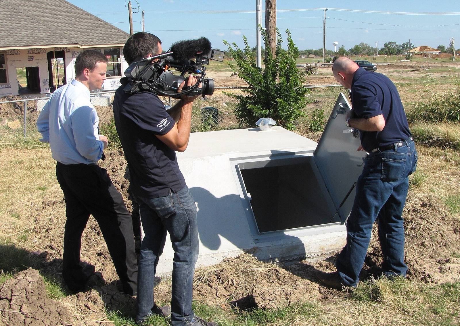 A FEMA mitigation expert opening the steel door of a partially underground concrete safe room beside a rebuilt house in Moore, Oklahoma, while a television crew films