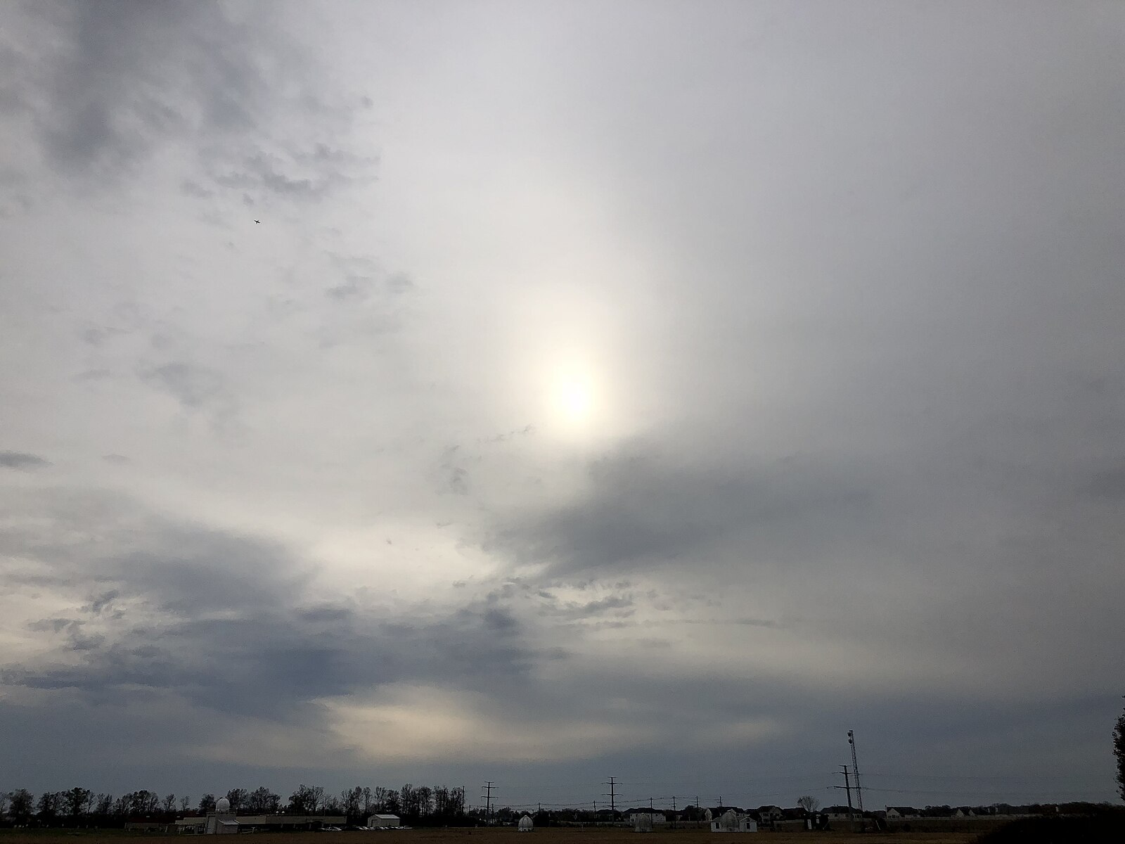 Sun visible as a bright disc through a thin, even layer of cirrostratus cloud over a flat landscape