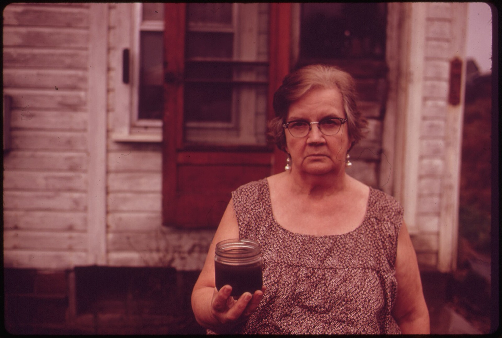 Mary Workman standing outside her Ohio farmhouse in 1973, holding a jar of dark, undrinkable water from her well