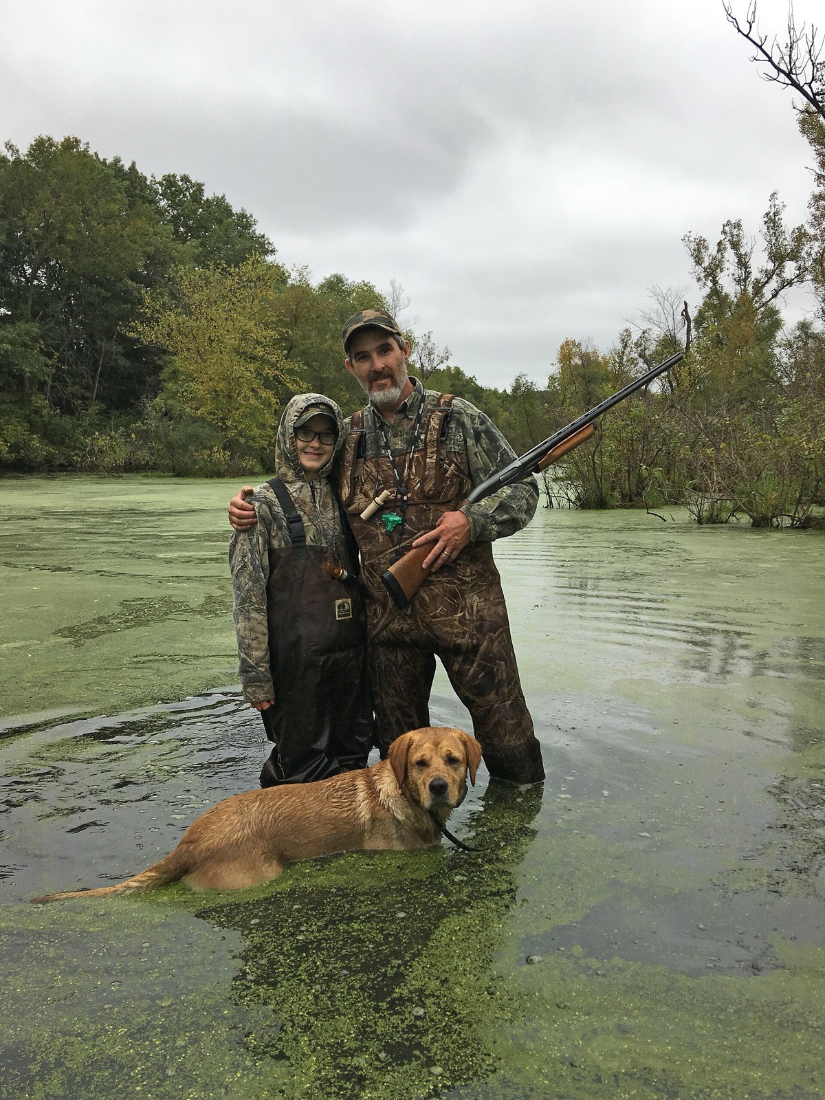 Father and daughter in waders with a retriever in a duckweed marsh