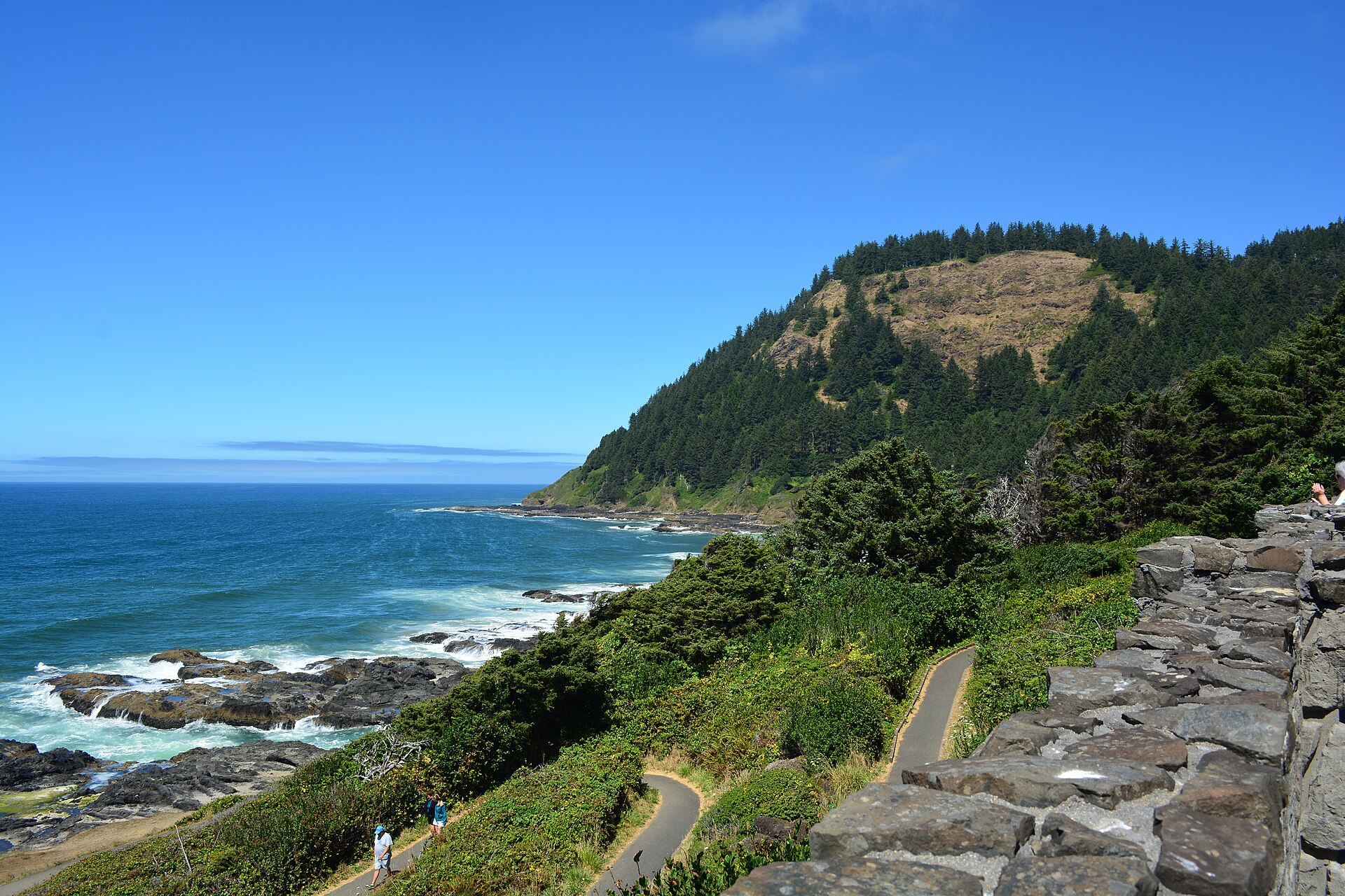 Elevated view north along the rocky Oregon coast from a stone-walled roadside overlook, with a paved path and two walkers below and a forested headland beyond