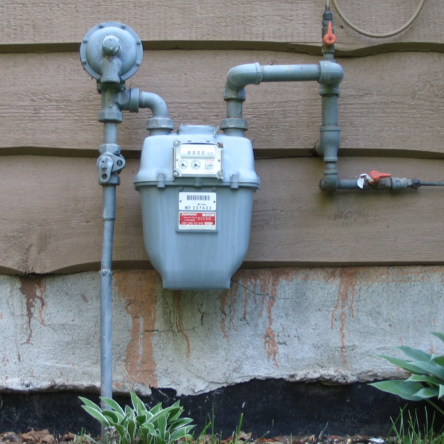 A residential natural gas meter with its regulator and shutoff valve, mounted against pale house siding