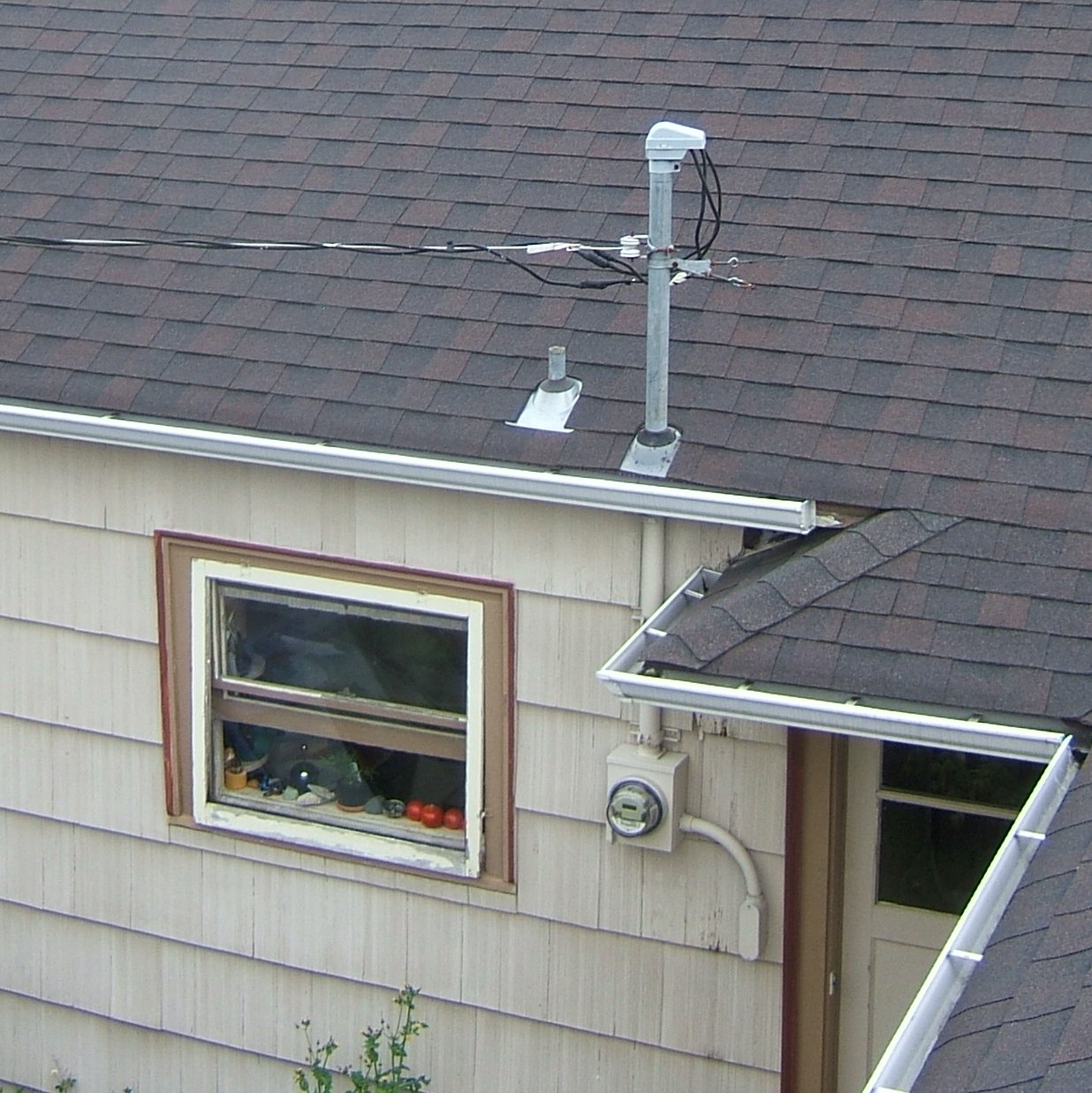 The corner of a shingled house showing the mast and weatherhead where overhead lines land on the roof, and the round electric meter mounted on the wall below