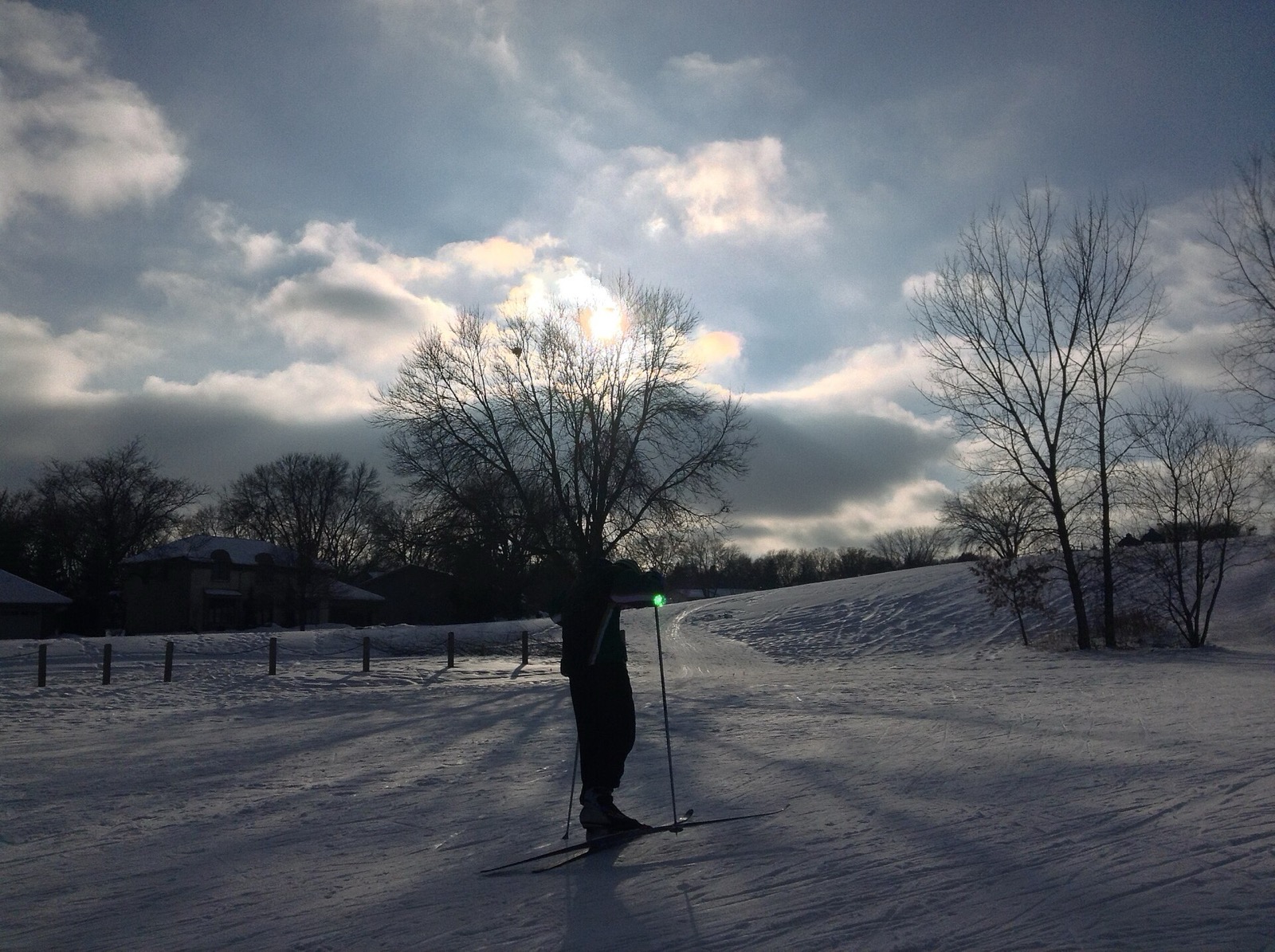 A skier silhouetted against a low winter sun on a groomed trail crossing open ground, with suburban rooftops and bare trees along the far edge