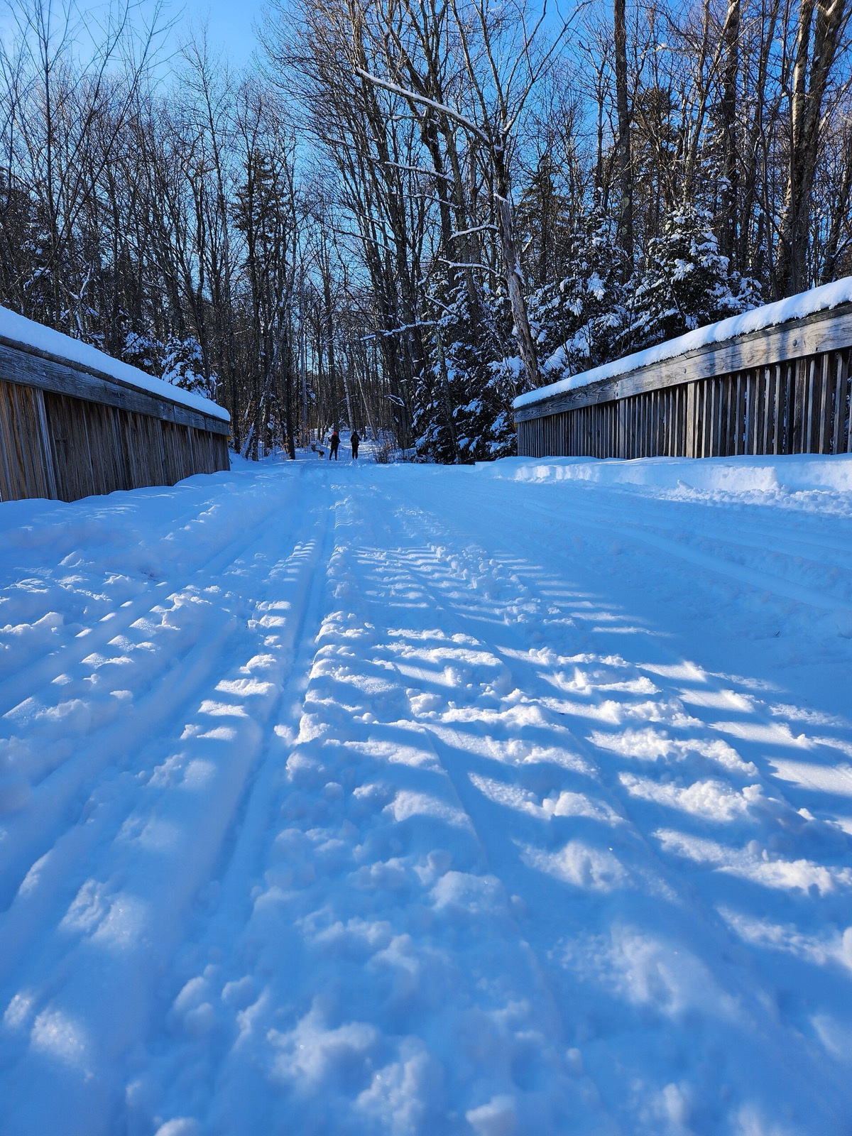 A low view along a snow-covered bridge deck between weathered wooden railings, the surface cut by overlapping ski tracks and boot prints, with two distant figures at the far end